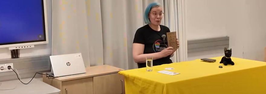 Roxane sitzt an einem Tisch mit gelber Tischdecke und hält das Buch Inepu hoch.